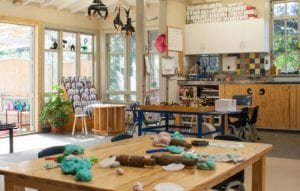 Sunlit PolyHigh playroom with playdough and rolling pins on a wooden table, a book display and doors open to the outdoor area